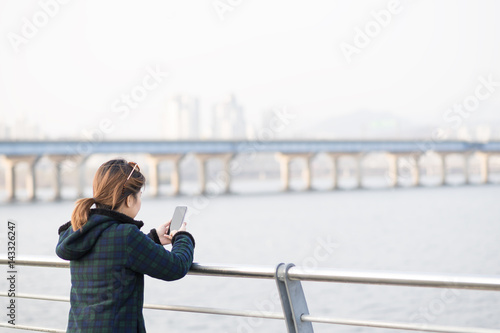 Woman waer fleece jacket standing and looking at smartphone in chilling day in evening at river side. Feeling lonely or chilling.