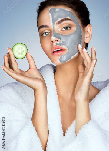 Beautiful sensual girl with the slice of cucumber in hand on blue background. Photo of girl in white bathrobe and with mask of clay on face. Grooming himself