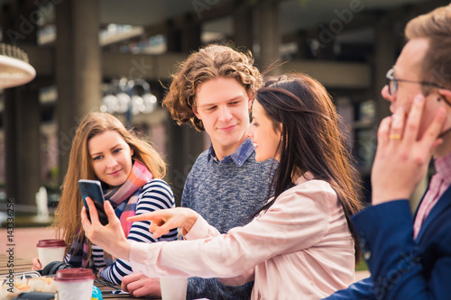 Happy clever students sit and look at phone which a woman shows in time of a break, outdoors