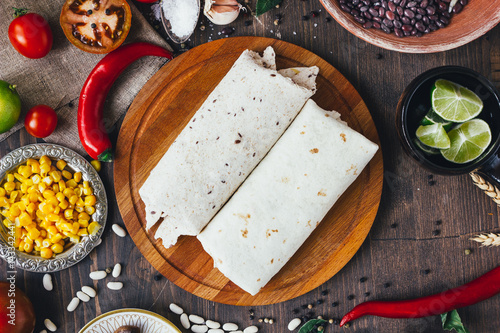 Vegetarian burrito on wooden board over black table surrounded by ingredients.