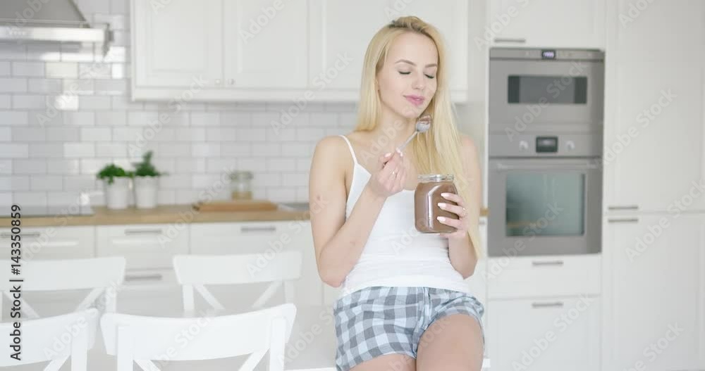 Young beautiful blonde sitting on table in modern kitchen and enjoying chocolate spread from jar with spoon. Young beautiful blonde sitting on table in modern kitchen and enjoying chocolate spread from jar with spoon.
