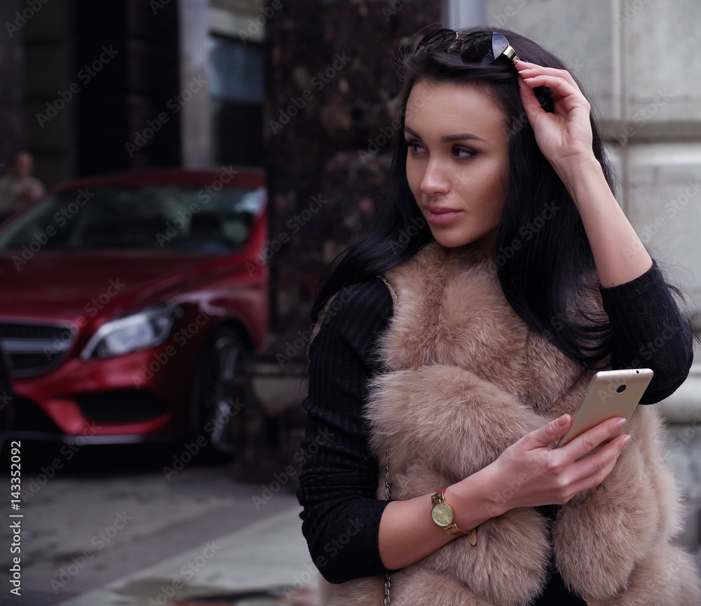 Beautiful young woman in fur vest with phone in hand on background of modern building and red car
