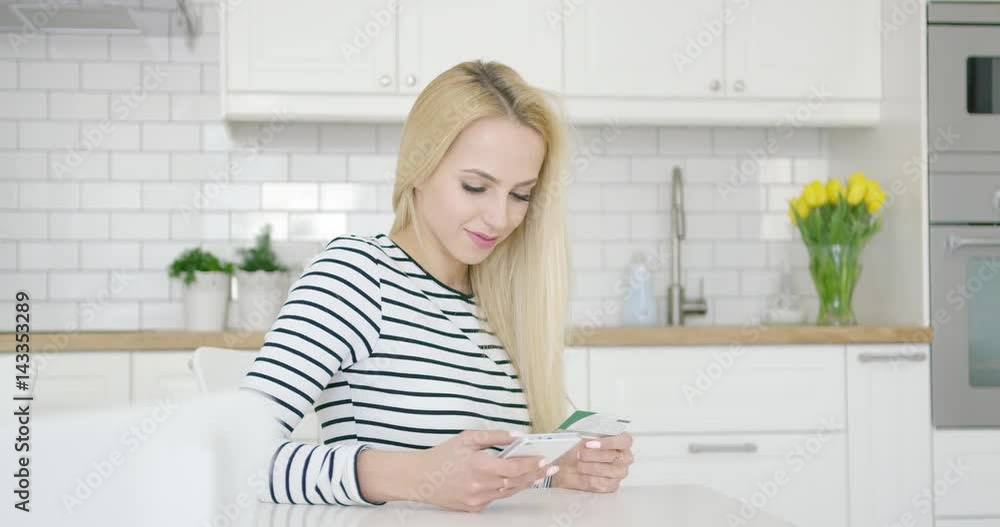 Smiling woman using credit card and phone to buy in Internet while sitting at table in kitchen. Smiling woman using credit card and phone to buy in Internet while sitting at table in kitchen.