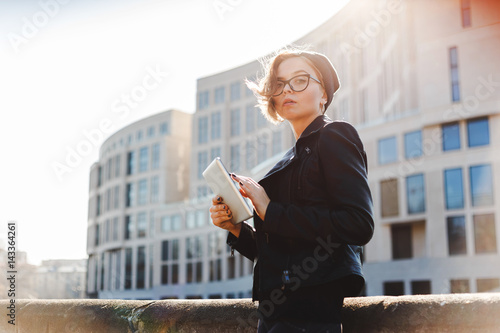 Portrait of a young woman with glasses, a knitted hat and a black leather jacket with an electronic tablet. Street photos. photo