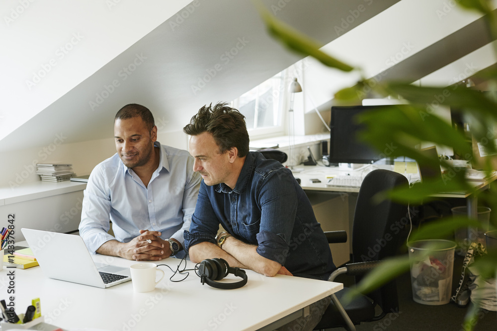 Sweden, Businessmen sitting side by side and looking on laptop in office Sweden, Businessmen sitting side by side and looking on laptop in office