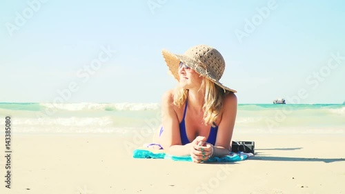 junge frau im blauen bikini liegt am strand mit einer vintage kamera und hat einen sonnenhut auf