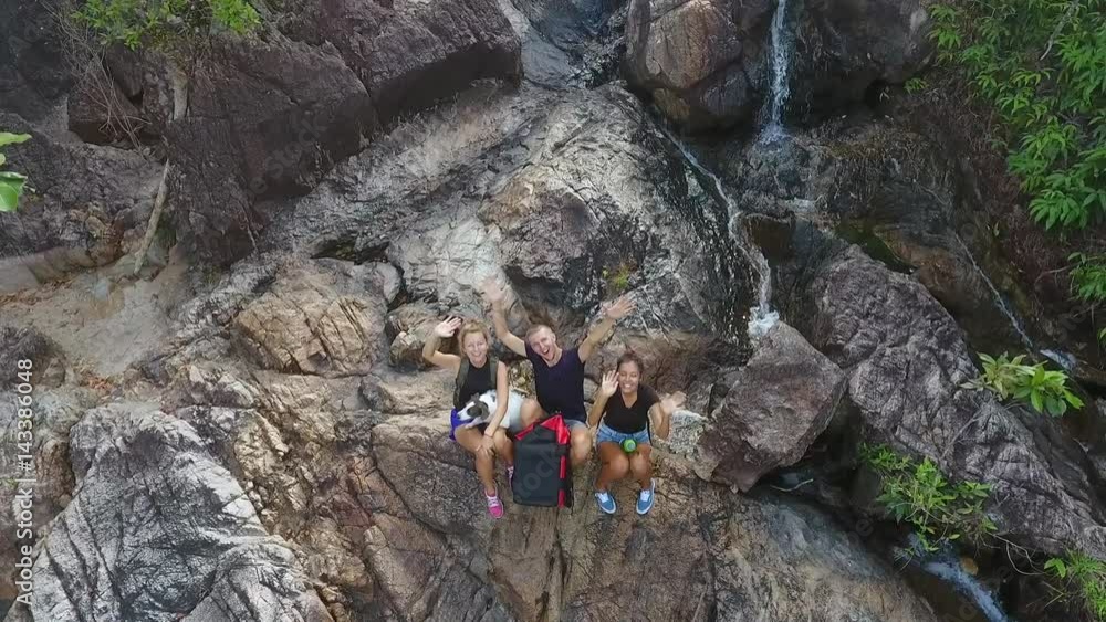 Aerial: Group of Young Tourists Resting at the Cascade Waterfall and Waving Their Hands in the Camera. Thailand. Aerial: Group of Young Tourists Resting at the Cascade Waterfall and Waving Their Hands in the Camera. Thailand.