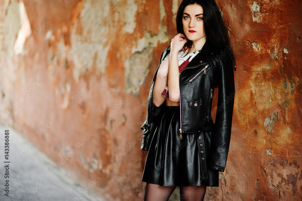 Young goth girl on black leather skirt and jacket against grunge wall.