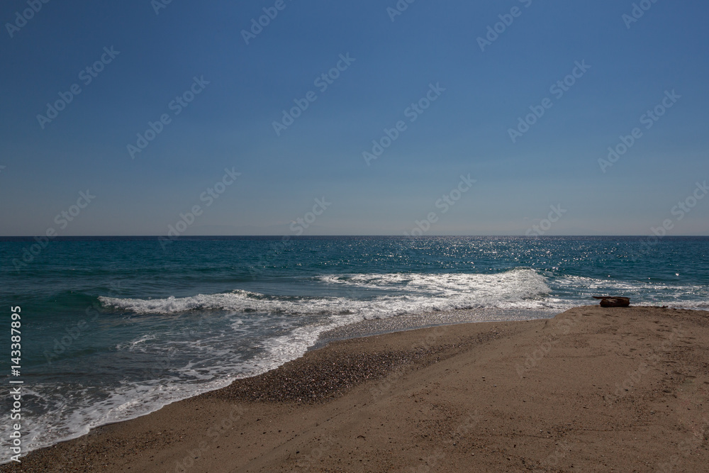 The surf of the blue turquoise sea with white perpendicular waves on the sandy beach against a bright clear sky.