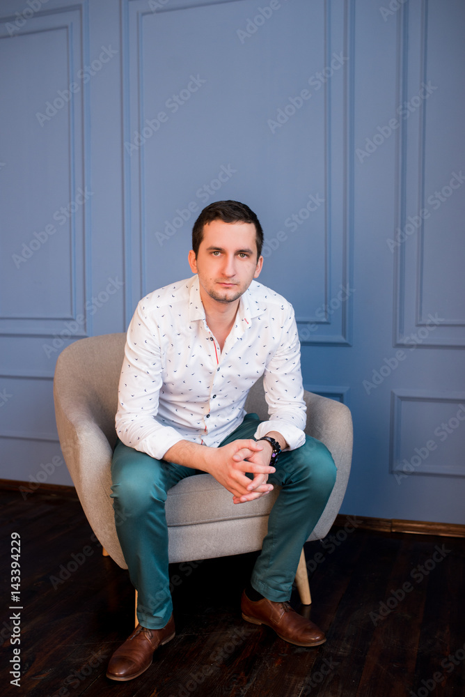 Happy handsome young businessman in white shirt and green trousers sitting in armchair and looking at camera Happy handsome young businessman in white shirt and green trousers sitting in armchair and looking at camera