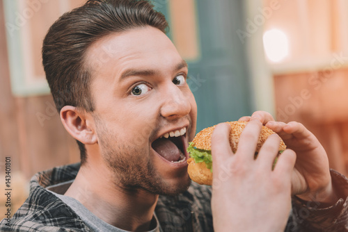 Excited young man eating fresh tasty hamburger and looking at camera