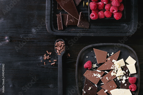 Variety of dark, milk and white chopping chocolate on old plate and spoon with fresh raspberries in black wood box over black burnt wooden background. Top view with space. Chocolate dessert concept