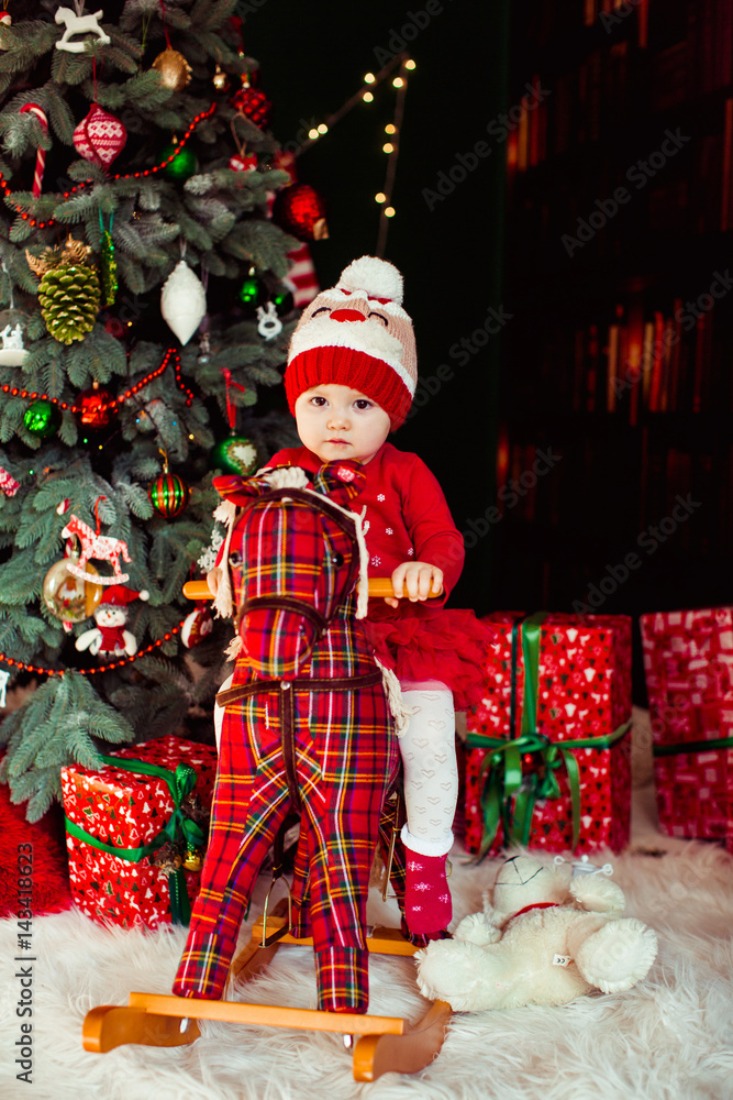 Little child in warm hat sits on toy horse before Christmas tree