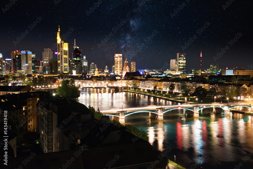 illuminated skyline of Frankfurt am Main downtown financial district seen from the east with river in foreground at night with sky full of stars