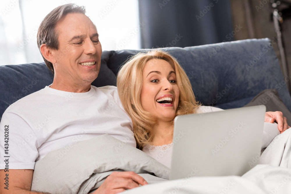 Happy middle aged couple using laptop and lying on bed at home Happy middle aged couple using laptop and lying on bed at home