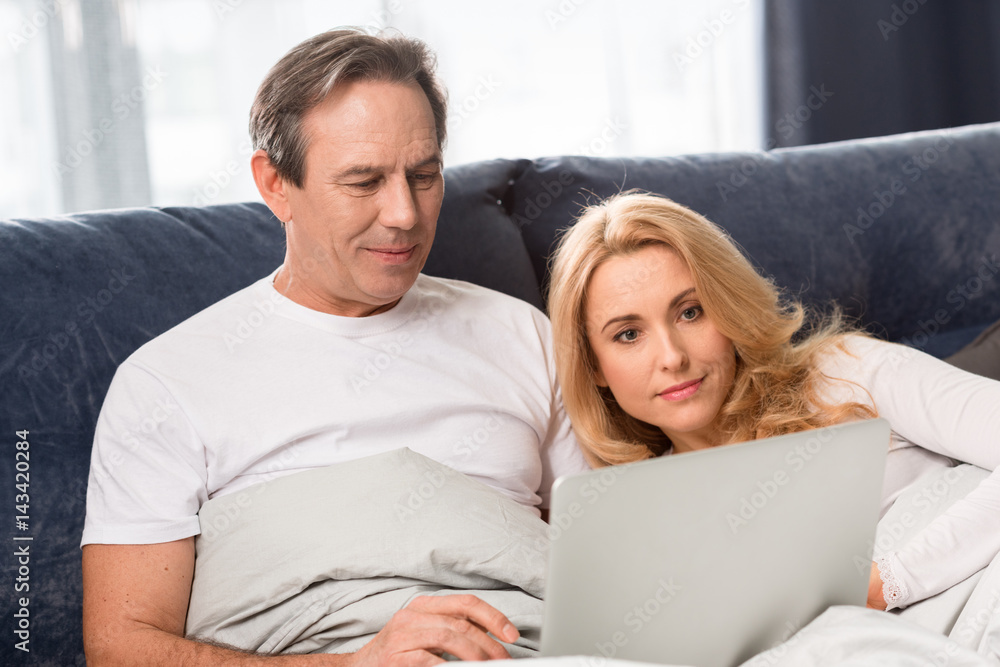 Happy middle aged couple using laptop and lying on bed at home Happy middle aged couple using laptop and lying on bed at home