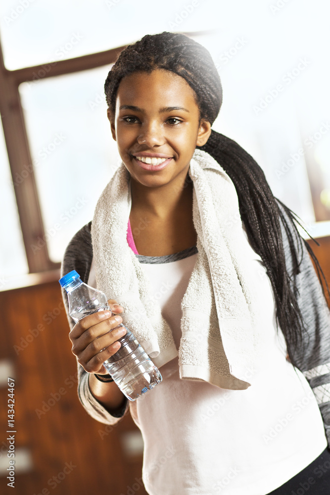 Beautiful young girl in a gym. Cute African girl on a training. She's  holding a water and smiling. Indoors. Stock Photo