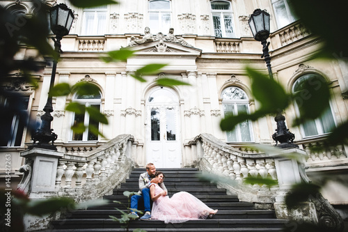 Look from behind green bushes at wedding couple sitting on the stone stairs to an old house