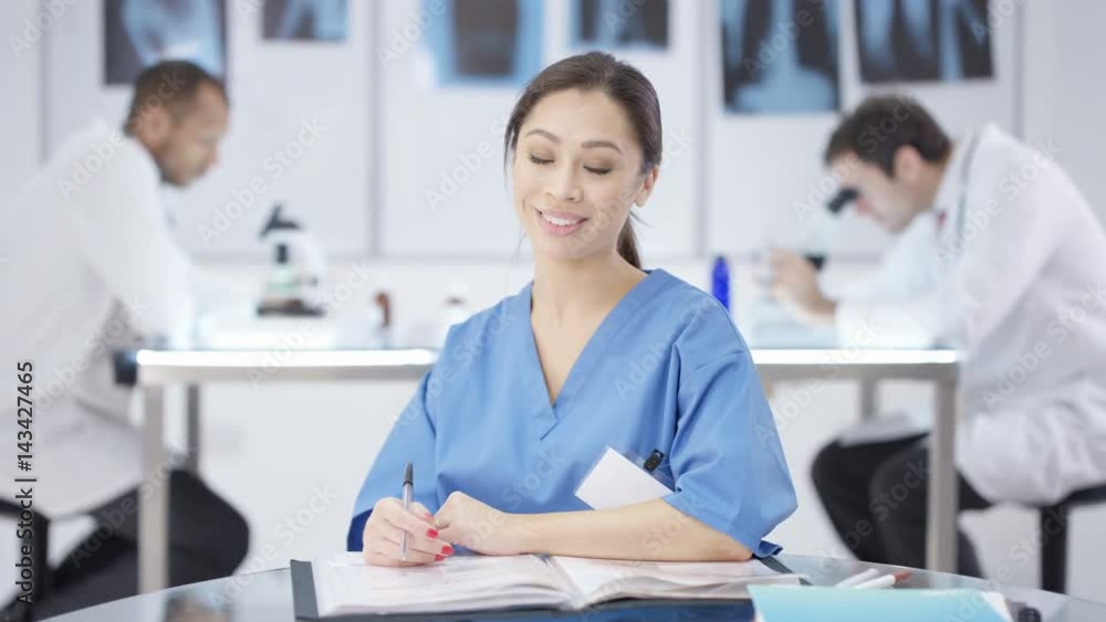 Portrait smiling doctor sitting at desk with colleagues working in background Portrait smiling doctor sitting at desk with colleagues working in background