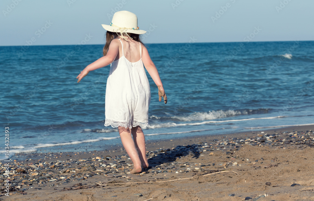 Little girl in white dress walking alone the sea, playing on the seashore at the sunset