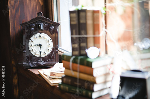 Old wooden clock stands behind bunch of books on the windowsill