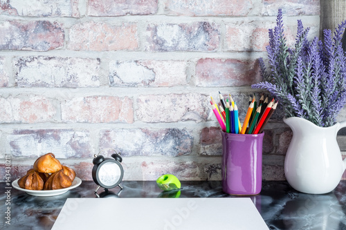 Colored pencils in a mug, vase of lavender flowers, clock, plate of cakes, white paper on a table against a brick vintage wall. photo