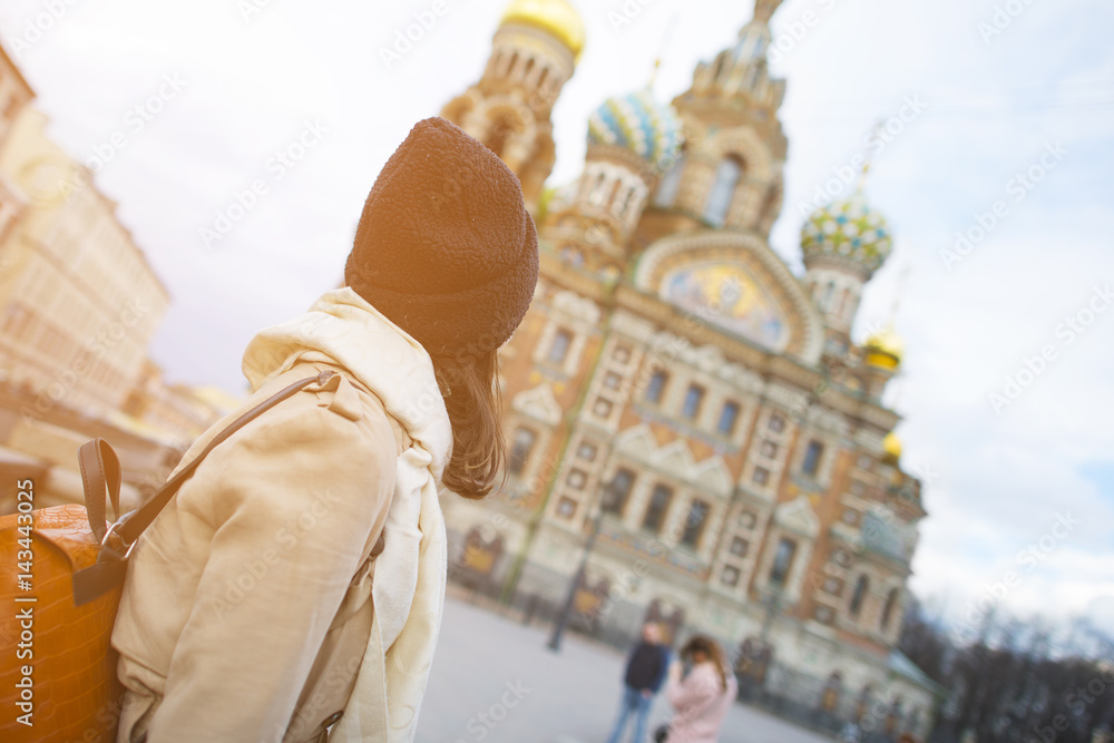 A woman hipster in a white coat and a black hat looks at the Church of the Savior on Blood in St. Petersburg, Russia.