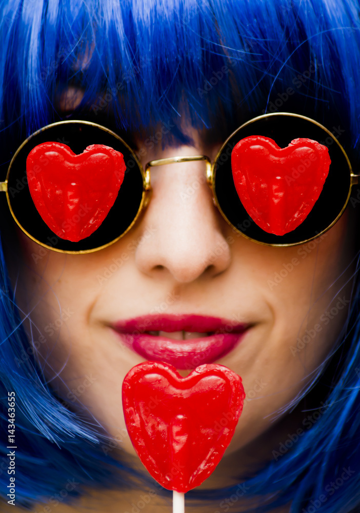 Beauty woman with blue wig and sunglasses and holding with her hand a caramel with heart shape, with the reflection of candy in glasses, portrait isolated on black background Beauty woman with blue wig and sunglasses and holding with her hand a caramel with heart shape, with the reflection of candy in glasses, portrait isolated on black background