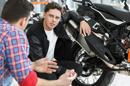 Tell me about this one. Portrait of a young handsome customer listening to motorcycle salon manager posing near a new motorbike © Nestor