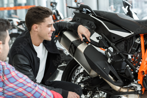 I love this one. Shot of a stunning hot handsome young man laughing cheerfully looking at the new motorbike while listening to motorcycle salon manager speaking