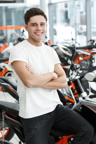 Excited today. Vertical shot of a smiling handsome biker man posing near his new motorcycle at the local salon © Nestor