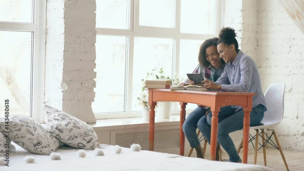Two attractive curly haired mixed race young girls sitting at the table have fun while learning lessons and using tablet at home Two attractive curly haired mixed race young girls sitting at the table have fun while learning lessons and using tablet at home