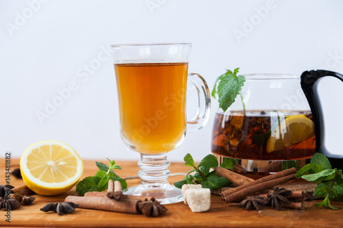 Clear big glass and teapot of hot black herbal tea with lemon, mint and Melissa leaves on light rustic wooden table. Summer, Autumn, winter drink. Cinnamon and cane sugar for decoration. 