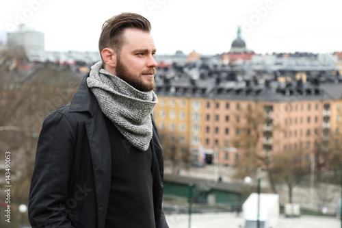 Lonely man wearing a grey scarf and black jacket, standing outside on a mountain with the city behind him on a cold cloudy day.