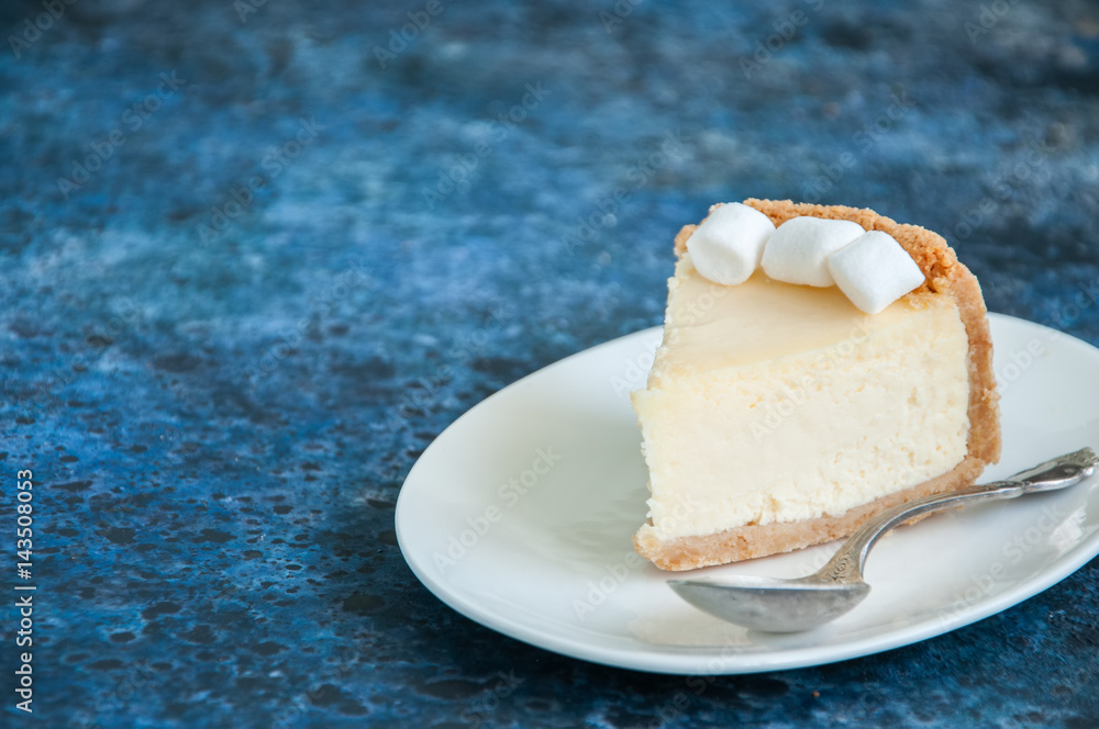 Slice of New York Cheesecake style with marshmallows on white plate on blue background Slice of New York Cheesecake style with marshmallows on white plate on blue background