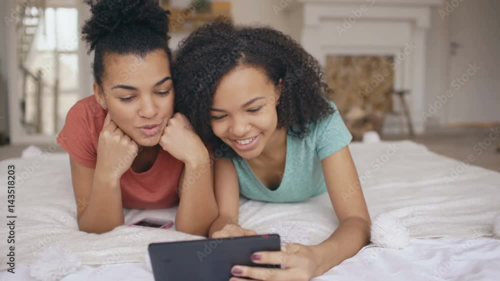 Two cheerful mixed race funny girlfriends sharing social media using tablet computer and talks lying in bed at home Two cheerful mixed race funny girlfriends sharing social media using tablet computer and talks lying in bed at home