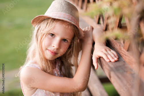 Summer portrait of cute blonde girl 5-6 year old wearing hat posing outdoors in park. Looking at camera. Childhood.