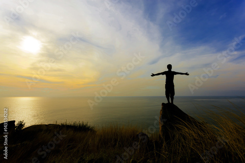Silhouette of a man with raised-up arms at the beautiful sunset  traveller man stands on a stone of sea stony coast.
