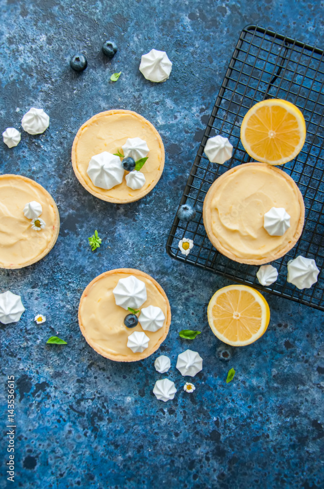 Fototapeta premium Individual lemon curd tarts with blueberry jam meringue basil leaves on a blue rusty background. Flat lay and copy space.