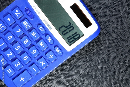 Close up photo of blue color solar and battery power calculator put beside the chrome ball pen on black leather surface background on calculator display panel represent number on screen.