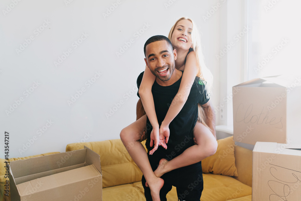 Young couple moving out, just moved in new house, looking at camera and smiling, enjoying relocating. Man lifting his woman on his back, labeled cardboard boxes around in room. Young couple moving out, just moved in new house, looking at camera and smiling, enjoying relocating. Man lifting his woman on his back, labeled cardboard boxes around in room.