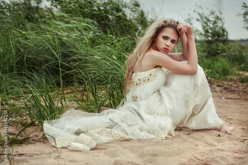 Beautiful girl in a white dress is sitting on the beach and looking back.