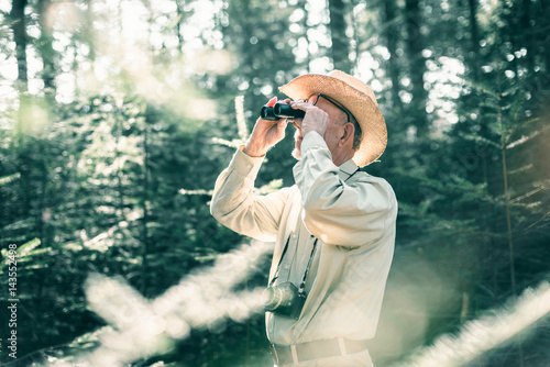 Man with hat and camera looking through binocular in forest.