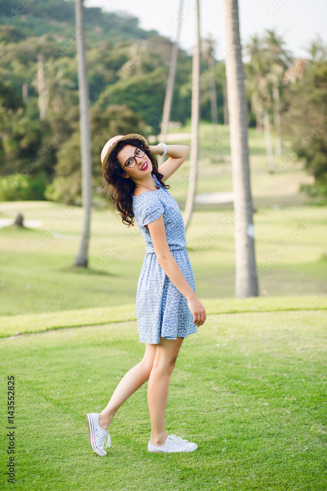 Young girl wearing sky-blue dress is standing in a tropical park. Girl has straw hat and black glasses on