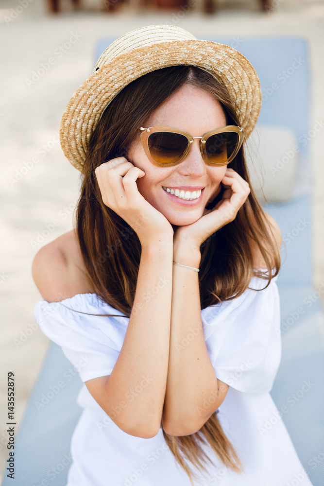 Cute girl in brown sunglasses and straw hat smiles widely and holds her hands near chin. She wears white dress with open shoulders. She sits on a beach chaise-longue.