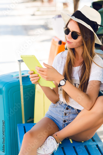 Cute girl sitting on a blue bench and playing on a tablet in yellow case. She wears denim shorts, white t-shirt, dark sunglasses, white sneakers and straw hat. She sits with two suitcases next to her.