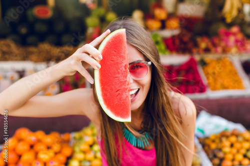 Closeup portrait of funny girl in pink sunglasses holding slice of watermelon on half face on tropical fruits background. photo