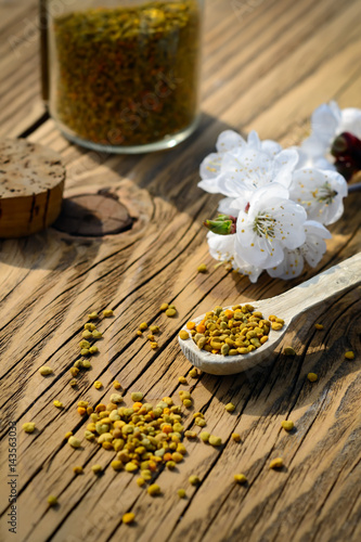 Grains of bee pollen in jar and wooden spoon on wooden table with flowers of spring trees. Apitherapy. Bee products.