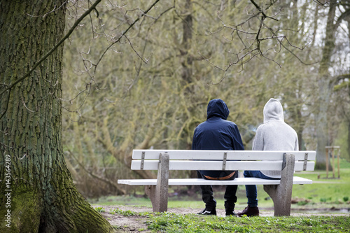 Two young men with hoods on a bench from behind in bad weather in the park, couple, homosexuals, refugees, homeless or just friends