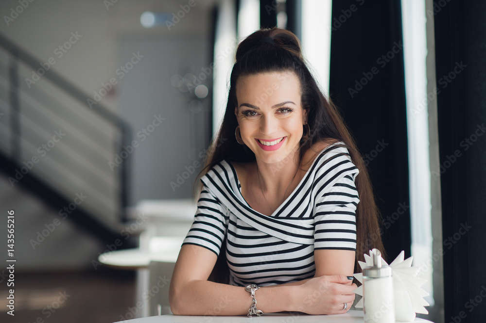 Young woman in striped blouse is sitting in cafe at wooden table near window and looking at the camera. Perfect smile, modern interior, indoor. Young woman in striped blouse is sitting in cafe at wooden table near window and looking at the camera. Perfect smile, modern interior, indoor.